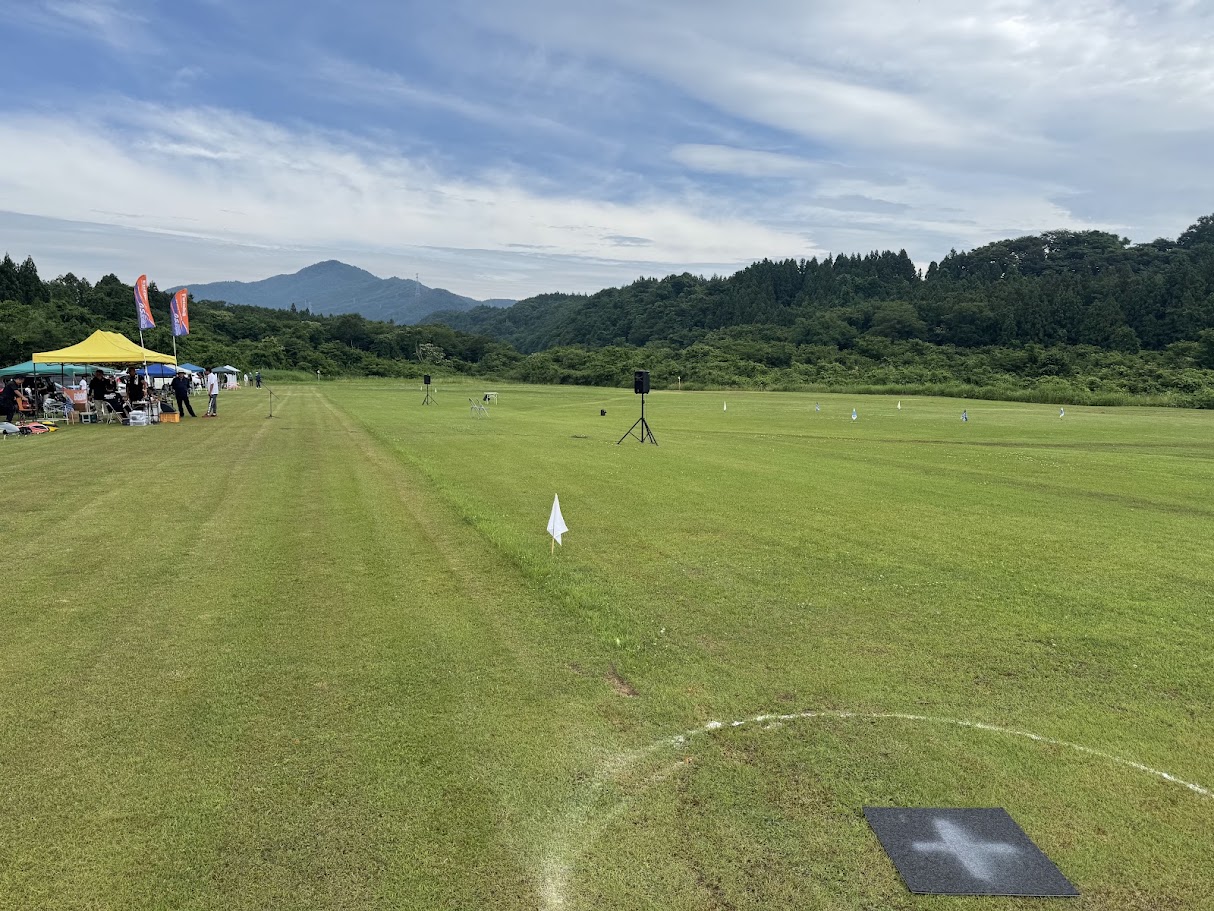 山に囲まれた広大な芝生のラジコン専用飛行場。大会のためのテントやのぼり旗、白線のサークルが見える